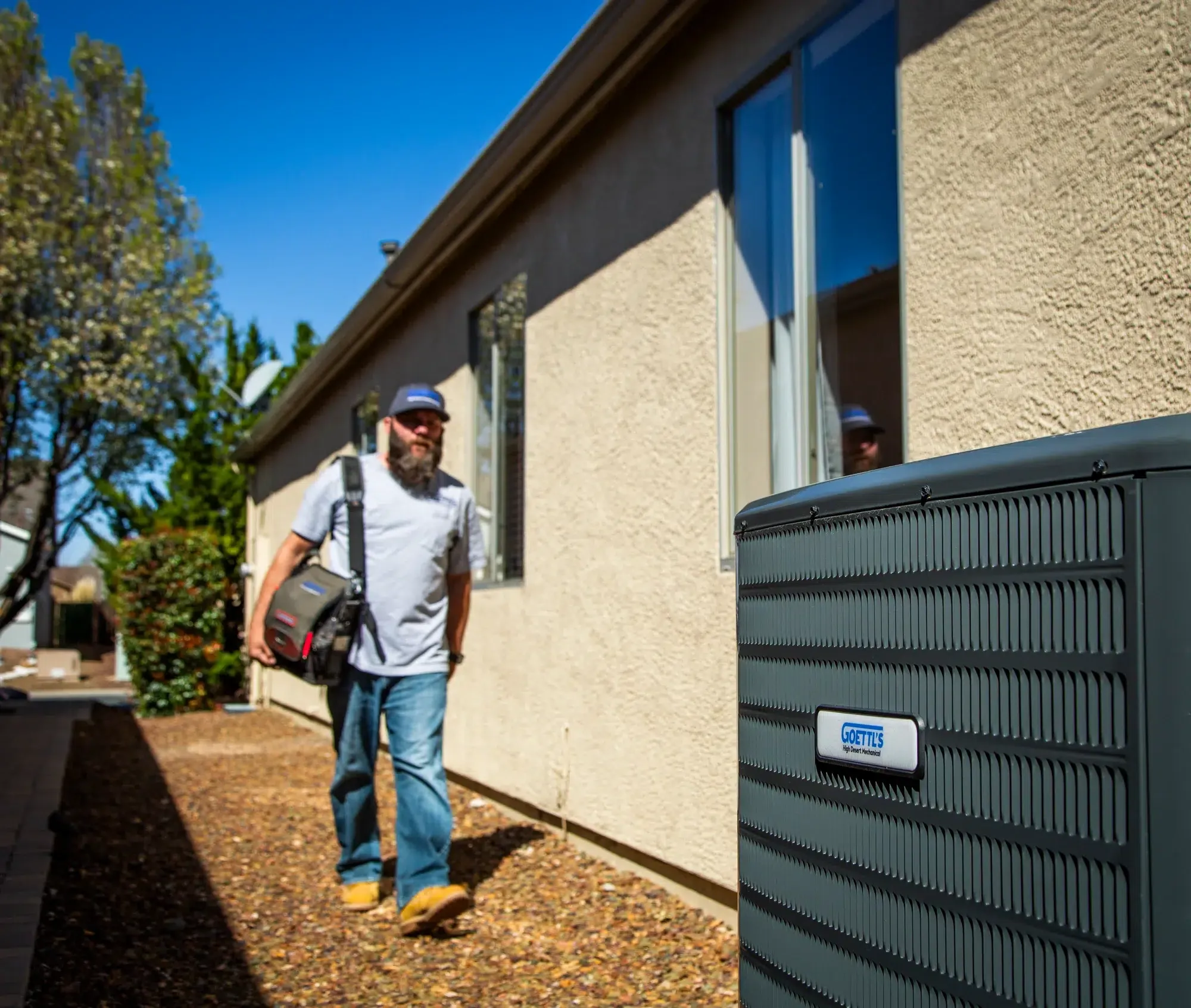 Goettl’s High Desert Mechanical technician approaching outdoor HVAC condenser beside home – Prescott Valley AZ
